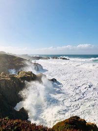 Scenic view of waves splashing on rocks at sea against sky