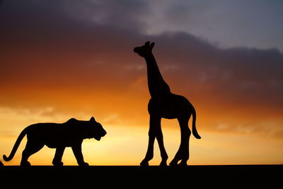 Close-up of silhouette tiger and giraffe toys against orange sky