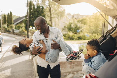 Boy looking at cheerful father carrying daughter by electric car on driveway