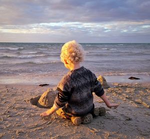 Rear view of boy sitting on shore at beach against sky