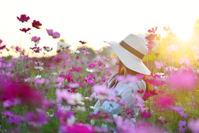 A woman in white dress holding white hat stand in  the purple, pink cosmos flowers field. 