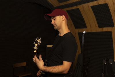 Caucasian man plays guitar, holding musical instrument in hands, standing in studio. darken photo
