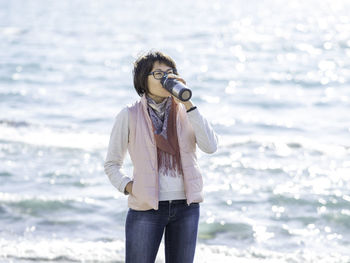 Woman drinks hot tea from metal travel mug. vacation on ocean coast. turquoise water behind woman.