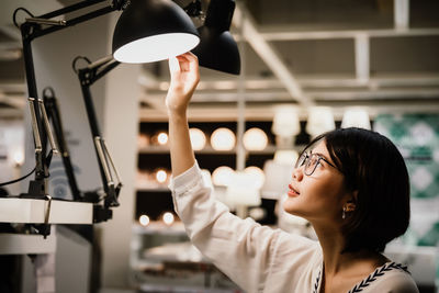 Asian young beautiful woman wearing glasses choosing new lamp to buy at furnishings lighting store