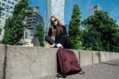 Young woman student, freelancer with backpack and laptop sitting on modern downtown high-rises