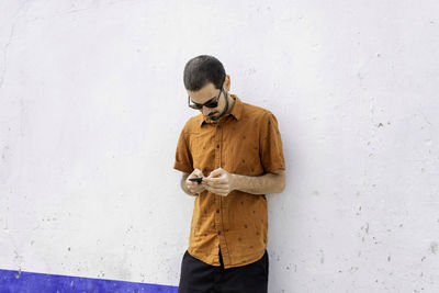 Young man using phone while standing against white wall