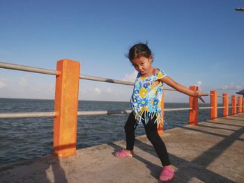 Girl making a face while standing by railing on pier over sea against clear blue sky