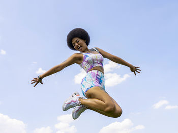 Low angle of delighted african american female in summer outfit in moment of jumping above ground on background of blue sky