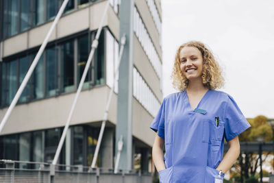 Portrait of happy female nurse with hands in pockets standing in front of hospital