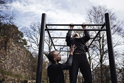 Side view of man assisting woman exercising on monkey bars in forest