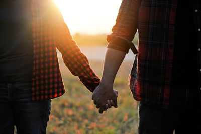 Midsection of couple holding hands while standing outdoors at sunset
