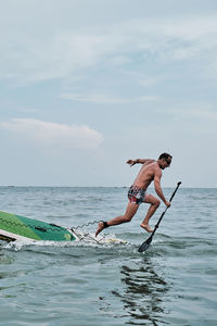 Full length of shirtless man on surfboard in sea against sky