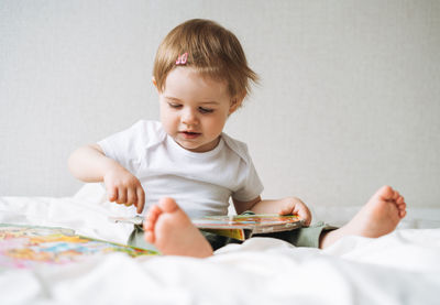 Cute funny baby girl in home clothes reading a book on bed at home