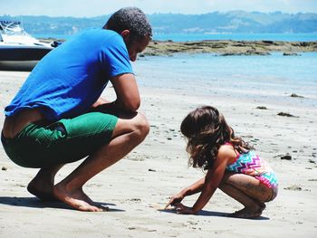 Side view of father looking at daughter drawing in sand at beach during sunny day