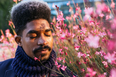 Carefree african american male standing in city park and enjoying aromatic smell of blooming pink flowers