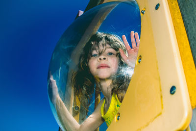 Low angle portrait of girl looking through glass against clear blue sky
