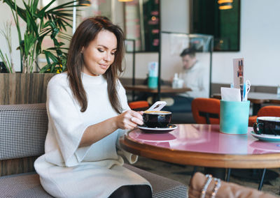 Attractive young brunette woman in white casual dress with cup of coffee using mobile phone in cafe