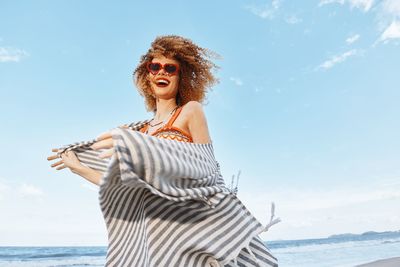 Portrait of young woman standing at beach against sky