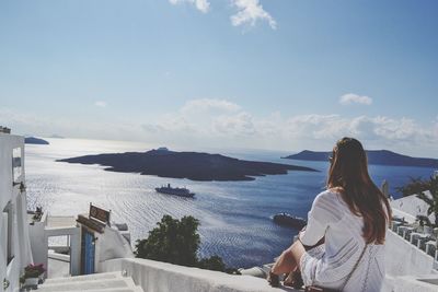 Rear view of woman sitting on retaining wall by sea against sky