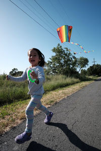 Cheerful girl holding kite running on road by grass and trees against clear sky