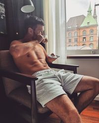 Shirtless man having tea while sitting on chair by window at home