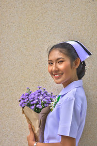 Smiling young nurse with bouquet standing against wall