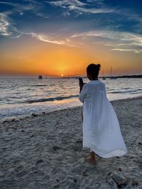 Rear view of woman standing at beach against sky during sunset