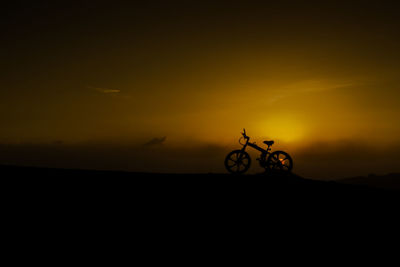 Silhouette person riding bicycle on field against sky during sunset