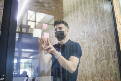 Male executive manager writing on sticky note on glass wall during brainstorm with coworkers in office