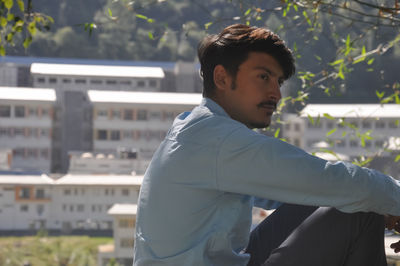 Close-up of a young guy looking sideways while sitting outdoor with buildings in background 