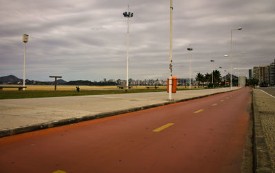 Road by street lights in park against cloudy sky