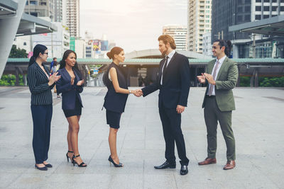 Colleagues clapping while coworkers handshaking on footpath against buildings in city