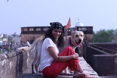 Portrait of smiling young woman sitting outdoors against sky with a labrador dog