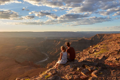 Rear view of couple sitting on rock against landscape and sky