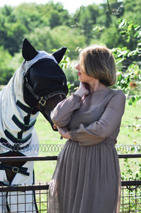 Blonde woman feeding a horse among tall trees in summer,countryside, rural