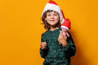 Cute girl in green dress and santa hat standing looking at camera with crochet toy during christmas celebration against yellow background