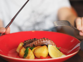 Close-up of hand holding knife and fork near red plate with octopus and potatoes dishes 