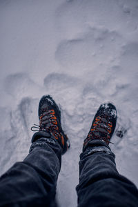 First person view on a man's legs with a black jeans and black and orange hiking boots on snow.