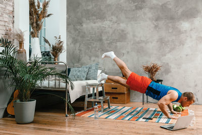 Cheerful sportsman with black hair does push-ups and watching online workout from laptop in bedroom