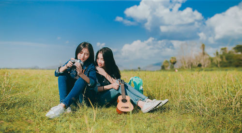 Young women sitting on grass while holding ukulele and camera against sky