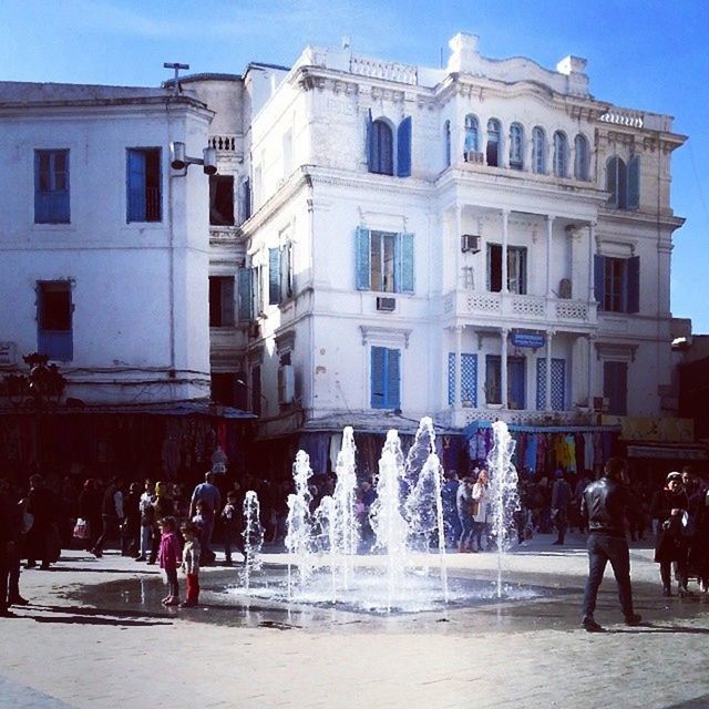 building exterior, architecture, built structure, large group of people, person, men, lifestyles, fountain, city, leisure activity, city life, motion, street, walking, building, sky, mixed age range, town square, spraying