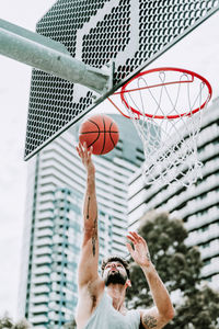 Determined young male basketball player in sportswear leaping and shooting ball near hoop while training alone on street playground