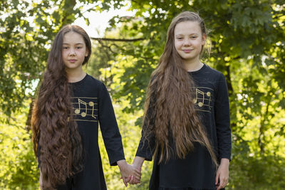 Portrait smiling two sisters with extra long hair holds hands in sunny day, girls wear black dresses