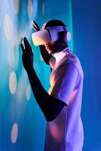 Side view of concentrated young african american in white t shirt and modern vr goggles touching screen while interacting with virtual object in dark studio with neon illumination