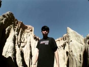 Portrait of young man standing on rock formation against clear blue sky