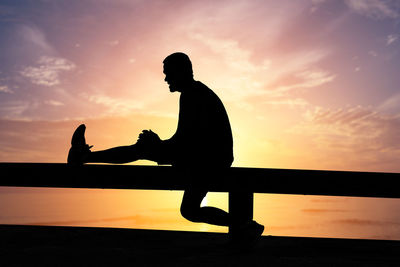 Silhouette man sitting on railing against sky during sunset