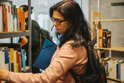 Side view of mature female student searching book on rack in college library