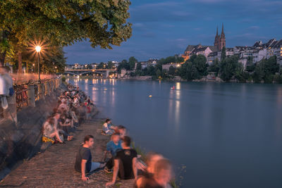 People on illuminated city by river against sky