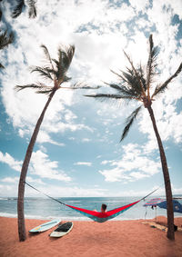 Man on hammock by palm trees at beach against sky