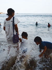 Playful boy splashing water on sister with mother standing on shore at beach